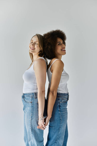 Две женщины, одна черная и одна белая, стоят спиной к спине, улыбаясь, в белых майках и синих джинсах.