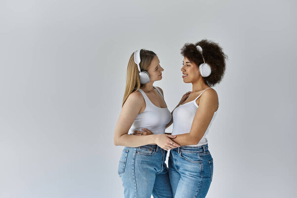 Two women in white tank tops and jeans stand facing each other, smiling, while wearing headphones.