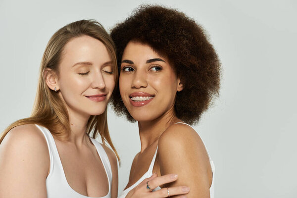 Two women, one blonde and one dark curly hair, wear white tanks and jeans, smiling at each other against a grey backdrop