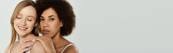 Two women in white tanks, one blond and one with dark curly hair, smiling and looking at the camera