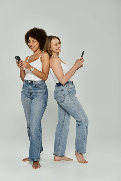 Две женщины в белых майках и джинсах стоят спиной к спине и улыбаются, глядя в свои телефоны.