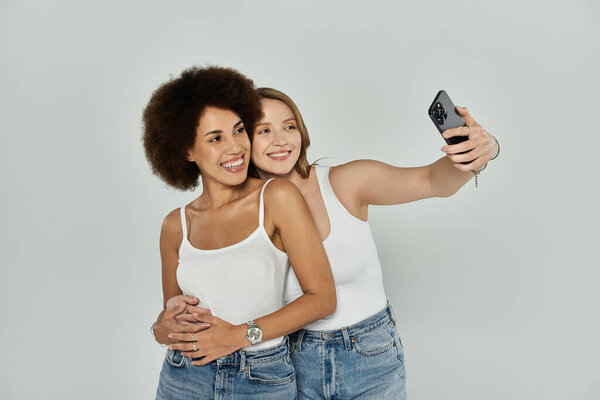 Two women, one with curly hair and one with straight hair, wearing white tank tops and jeans, pose for a selfie.