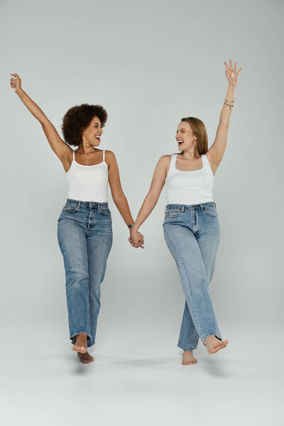 Две женщины в белых майках и джинсах идут рука об руку и смеются на белом фоне..