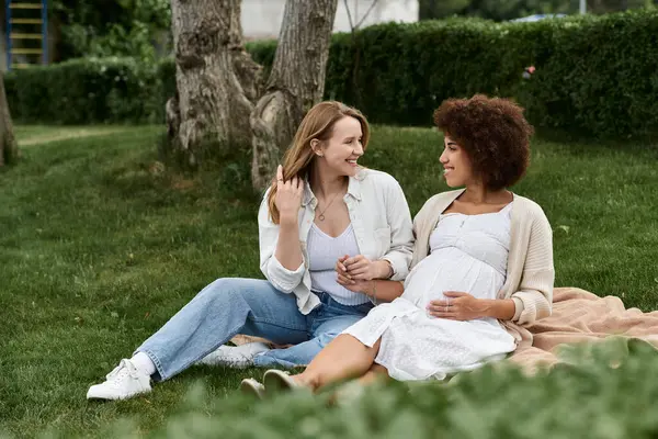 stock image Two women, one pregnant, sit on a blanket in a grassy park, smiling and looking at each other.