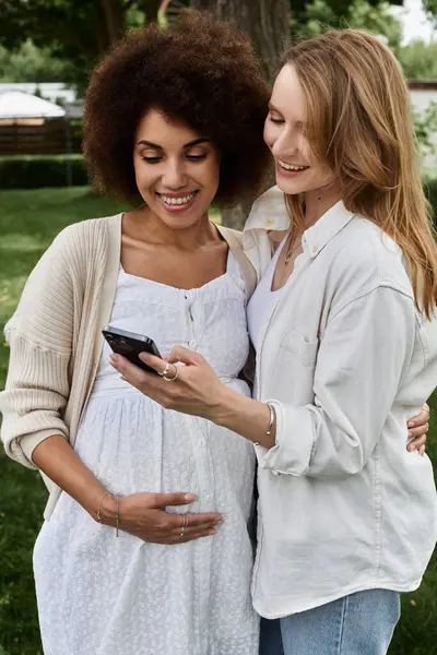 stock image Two women, one pregnant, stand in a park and look at a phone together. They are smiling and happy.