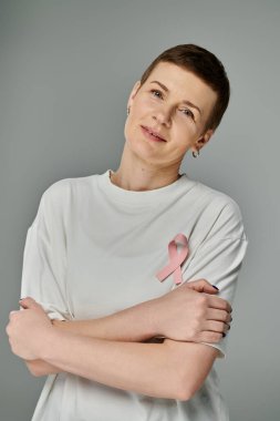 Kısa kahverengi saçlı, pembe kurdeleli beyaz tişörtlü bir kadın, kollarını gri bir arka planın önünde kavuşturuyor..