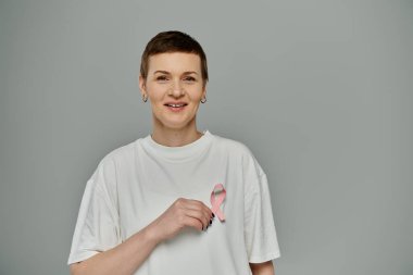Kısa saçlı bir kadın, beyaz gömleğinde pembe bir kurdele tutarken gülümsüyor ve meme kanseri farkındalığını savunuyor..