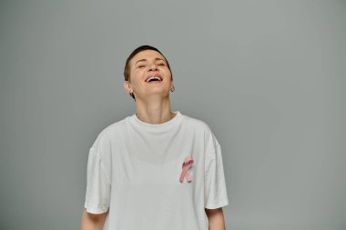 Kısa saçlı, günlük kıyafetler giyen ve pembe kurdeleli bir kadın gri bir arkaplanda parlak bir şekilde gülümsüyor..