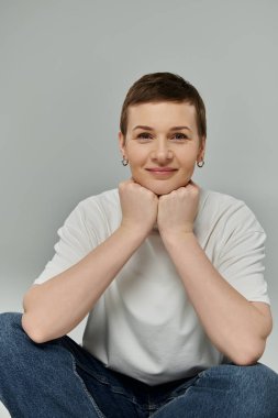 Kısa saçlı bir kadın elleri kucağında oturur, pembe kurdeleli beyaz bir gömlek giyer..