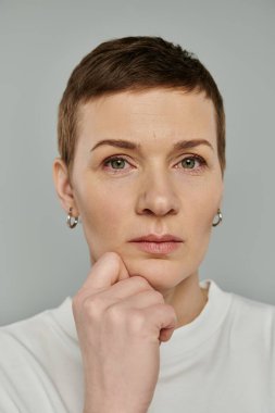 Kısa saçlı bir kadın, gri bir arka planda, yüzü düşünceli bir şekilde çenesini eline dayar..