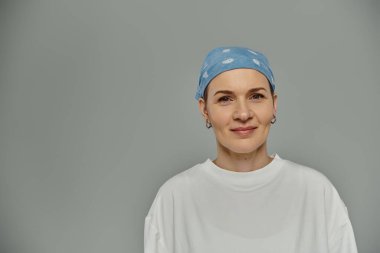 Kısa saçlı bir kadın mavi bir bandana takıyor ve yavaşça gülümsüyor, gri bir zemine karşı duruyor..