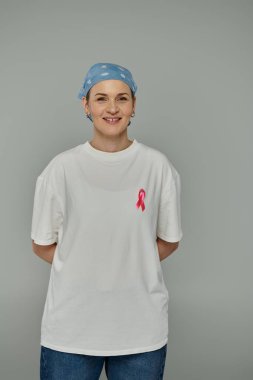 Kısa saçlı bir kadın mavi bir başörtüsü ve pembe kurdeleli beyaz bir tişört giyerken ışıl ışıl gülümsüyor..