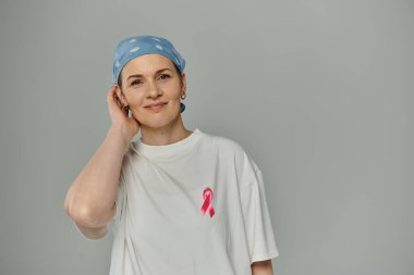 Kısa saçlı bir kadın beyaz gömleğine pembe kurdele takıyor, gri arka planda duruyor..