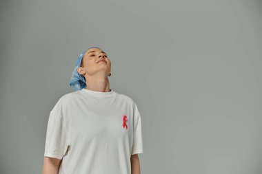 Kısa saçlı, beyaz tişörtlü ve mavi başörtülü bir kadın gömleğine pembe bir kurdele takmış yukarı bakıyor..