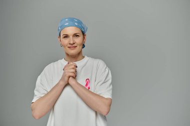 Kısa saçlı ve gömleğinde pembe kurdele olan bir kadın gri bir arka planda duruyor..