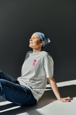 Mavi bandana takan ve gömleğinde pembe kurdele olan bir kadın gri bir arka planda oturur ve yukarıya bakar..