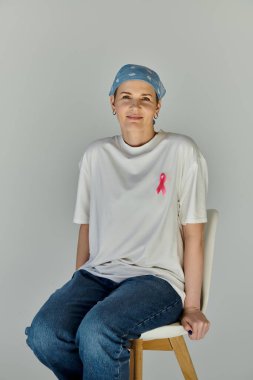 Sandalyeye oturan bir kadın pembe kurdeleli beyaz bir tişört giyiyor. Bu da meme kanseri farkındalığını gösteriyor..