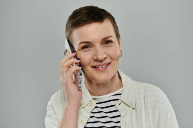 Kısa kahverengi saçlı bir kadın, gri arka planda akıllı bir telefonla konuşurken sıcak bir şekilde gülümsüyor..