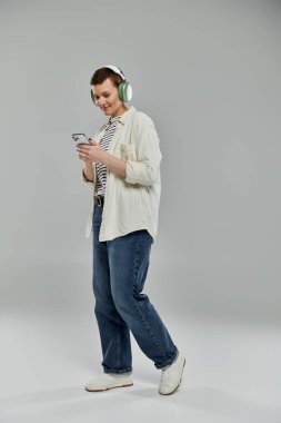 Kulaklık takan sıradan bir kadın gri bir arkaplanda dikilirken telefonunu kontrol ediyor..