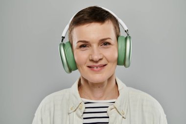 Kısa saçlı, günlük kıyafetler giyen bir kadın gülümserken kulaklıkla müzik dinliyor. Gri bir geçmişi var..