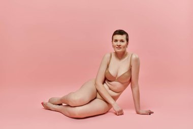 A woman with short hair wearing a bra sits on a pink background, posing for breast cancer awareness.