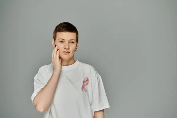stock image A woman with short hair and white t-shirt stands against a grey backdrop, hand on cheek, in a thoughtful pose