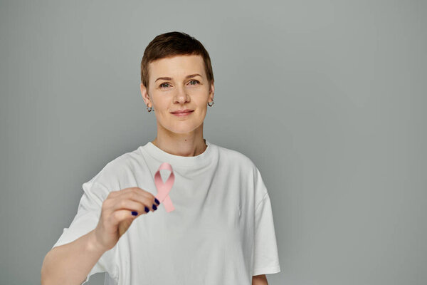 A woman with short hair, wearing casual attire, holds a pink ribbon in front of a grey background.