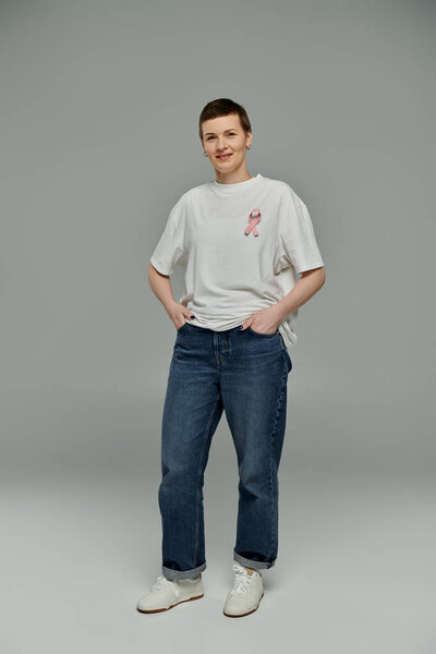 Женщина с короткими волосами, одетая в повседневную одежду и розовую ленту, уверенно стоит на сером фоне.