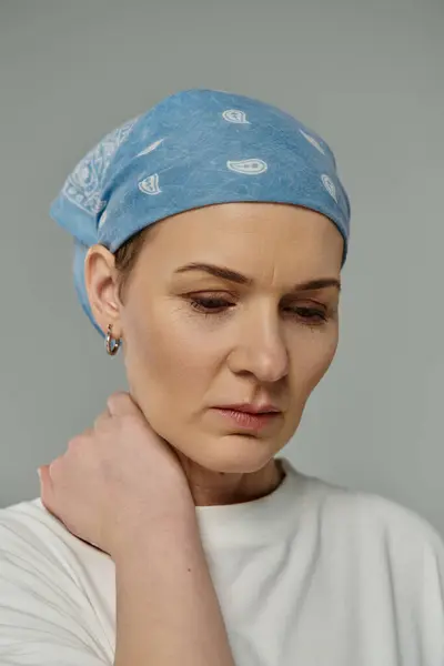 stock image A woman with short hair and a blue bandana rests her hand on her neck, lost in thought against a grey background.