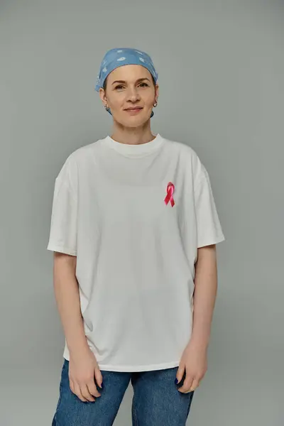 stock image A woman with short hair, wearing a white t-shirt and blue jeans, stands against a grey background with a pink ribbon pinned to her shirt.
