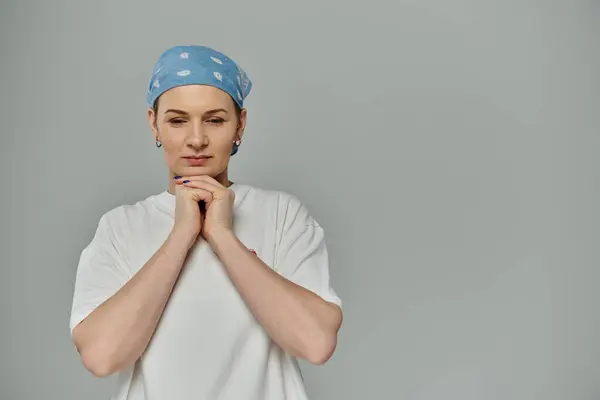 stock image Short-haired woman in white t-shirt, blue bandana, against grey background, hands together, pink ribbon on shirt