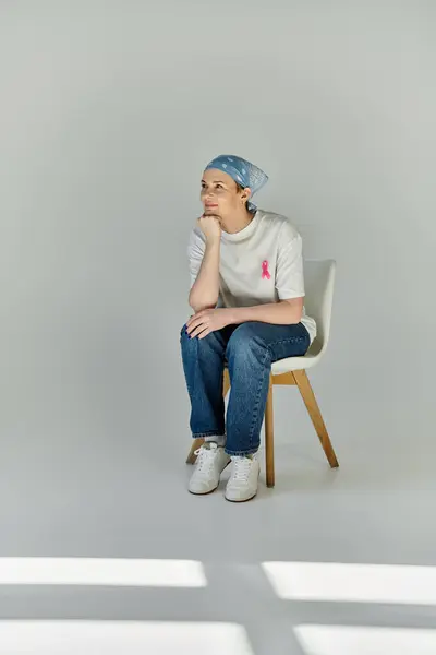 stock image A woman in casual attire sits on a chair, wearing a pink ribbon, in a studio setting.