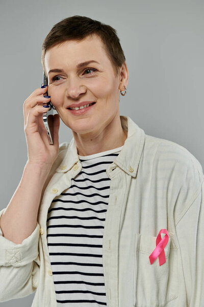 A woman with short hair smiles as she talks on the phone, wearing a white shirt with a pink ribbon for breast cancer awareness.