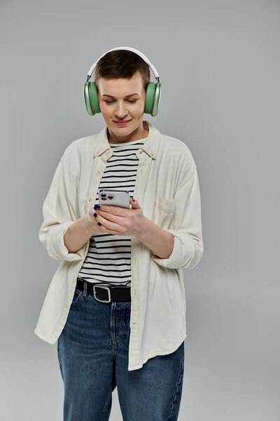 Женщина в наушниках и белой рубашке использует свой телефон, стоя на сером фоне.