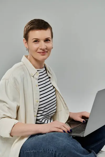 stock image A woman with short hair in casual attire smiles as she uses a laptop.