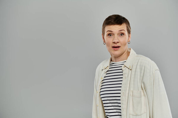 A woman with short hair is surprised and looks to the left of the frame. She is wearing casual attire and standing against a grey backdrop.