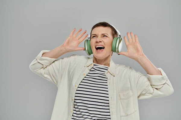 Short-haired woman in white shirt and striped tee dances, sings to music with headphones on gray background