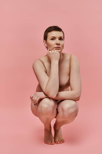 Женщина с короткими волосами, в лифчике, сидит в положении приседания на розовом фоне.