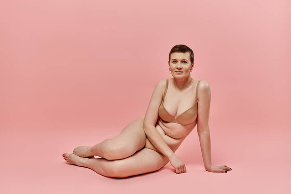 Женщина с короткими волосами в лифчике сидит на розовом фоне, позируя для информированности о раке молочной железы.