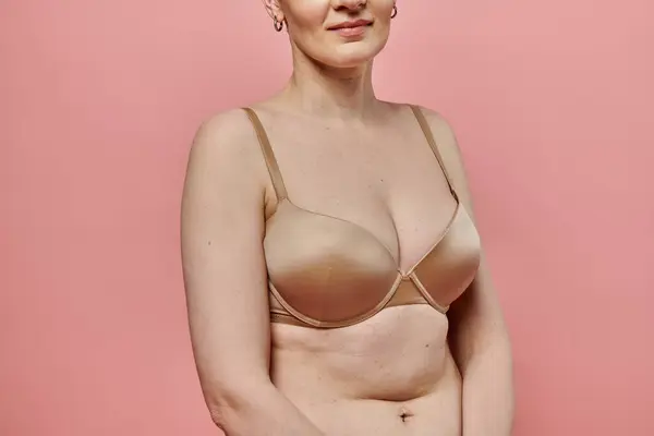 Stock image A woman stands against a pink backdrop, wearing a bra, promoting breast cancer awareness and prevention.