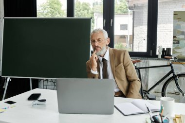 Sakallı bir adam elinde dizüstü bilgisayarla masada oturuyor, online bir derse hazırlanıyor, lgbtq öğretmeni.