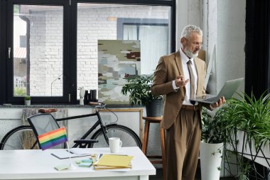 Orta yaşlı, sakallı, takım elbiseli ve kravatlı bir adam online ders vermek için dizüstü bilgisayar kullanıyor. Bir gökkuşağı bayrağı masayı süslüyor.