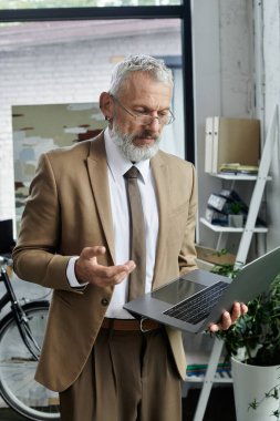 A middle-aged man with a beard, wearing a suit, teaches online through a laptop in a bright, modern office. clipart