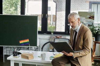 Orta yaşlı, sakallı ve gözlüklü bir adam, bir sınıfta online öğretmenlik yapıyor, bir masanın üzerinde belirgin bir gökkuşağı bayrağı sergileniyor..
