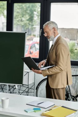 Daha yaşlı ve sakallı bir öğretmen modern bir sınıfta dizüstü bilgisayarını kullanarak online ders veriyor..