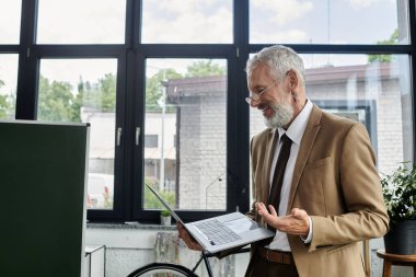 Orta yaşlı, sakallı bir adam ofisinden online sınıfa liderlik ederken gülümsüyor..