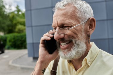 Gözlüklü sakallı bir adam modern bir binanın önünde cep telefonuyla konuşurken gülümsüyor..