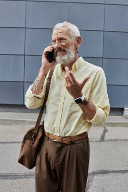 Sakallı, gözlüklü ve kolunda dövme olan olgun bir adam modern bir binanın dışında telefonla konuşuyor..
