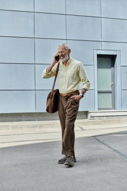 Orta yaşlı, sakallı bir adam modern bir binanın önünde telefonuyla konuşurken gülümsüyor..