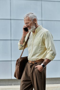 Orta yaşlı, sakallı ve gözlüklü bir eşcinsel modern bir binanın önünden geçerken cep telefonuyla konuşuyor..